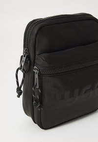 Černá nylonová crossbody taška s dvojitými zipy, texturovaným povrchem a výrazným vzorem loga "HUGO" na boku. Kompaktní a elegantní design.