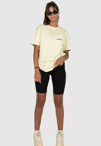 Pale yellow cotton t-shirt with a small black logo on the chest, paired with black bike shorts and high-top sneakers.