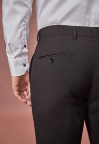 Pantaloni negri din stofă fină, cu un buzunar posterior cu nasture și o talie plată, asortați cu o cămașă de dress de culoare deschisă.