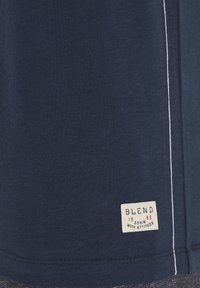 Marineblå bomulds t-shirt med hvid syning langs siden. Inkluderer et cremefarvet mærke med teksten "BLEND" og året "1993."