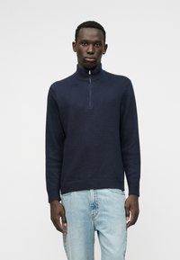 Maglione lavorato a maglia blu navy con colletto alto e zip a un quarto, caratterizzato da un motivo testurizzato e orli e polsini a costine. Abbinato a jeans azzurri chiari.
