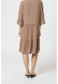 Vestido en capas de tejido marrón claro con manchas negras, con mangas tres cuartos y un dobladillo con volantes, combinado con zapatos negros de punta abierta.