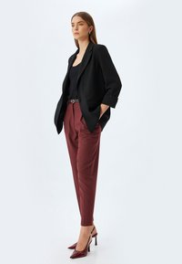 Schwarzer taillierter Blazer über einem schwarzen Top, kombiniert mit hoch geschnittenen, burgunderfarbenen Hosen und burgunderfarbenen spitzen Absätzen. Glattes Material, strukturierter Schnitt.
