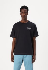 Black cotton t-shirt with a relaxed fit, featuring white embroidered text "VOLCOM STORE" on the chest and contrasting stitching details.