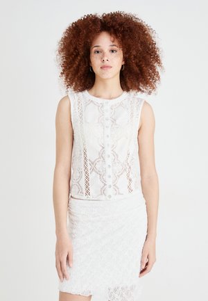 Witte mouwloze top met ingewikkelde kantdetails en knopen aan de voorkant, gecombineerd met een witte kanten rok met een scalloped zoom.