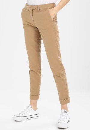 Persoon draagt opgerolde beige broek en witte lage sneakers met veters, staat met één hand in de zak tegen een witte achtergrond.