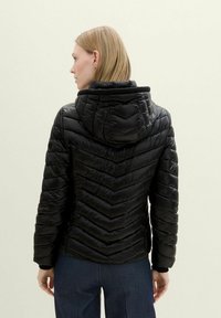 Chaqueta de plumas negra con un patrón de acolchado en forma de chevrón, capucha y puños elásticos. Textura suave y diseño ajustado, llevada sobre pantalones oscuros.