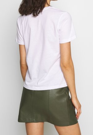 Kobieta ubrana w prosty biały t-shirt z krótkim rękawem oraz oliwkowozieloną skórzaną mini spódniczkę, widziana od tyłu.