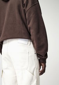 Sweat-shirt brun oversize à manches longues, associé à un pantalon en toile blanc avec une poche arrière et une étiquette blanche sur la ceinture.