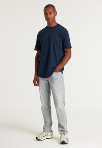 T-shirt in cotone blu navy con maniche corte e un piccolo logo nero sul petto, abbinato a jeans grigi chiari e sneakers beige.
