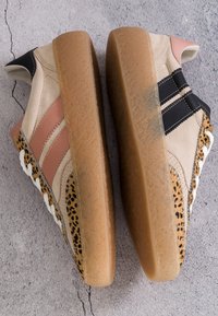 Paire de baskets beiges et marron avec imprimé léopard et rayures noires et roses, montrées semelle vers le haut sur une surface en béton fissurée.