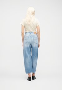 Άτομο με μακριά ξανθά μαλλιά, ντυμένο με μπεζ μπλουζάκι, φαρδιά ξεβαμμένα μπλε τζιν παντελόνια και μαύρα παπούτσια με τακούνι, γυρισμένο πλάτη προς την κάμερα.