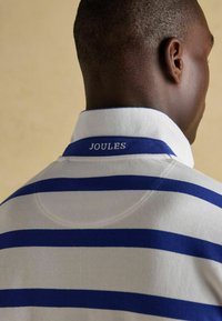 Valge polo särk siniste triipudega, millel on krae ja "JOULES" silt tagaküljel. Kangas näeb välja tekstuurne ja kerge.