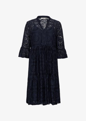 Frau lächelnd, trägt ein knielanges marineblaues Spitzenkleid mit dreiviertel Ärmeln, schwarze Sandalen mit Absatz und ein goldenes Armband.