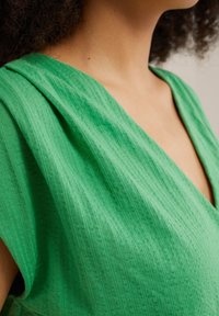 Abito verde con un tessuto liscio e strutturato, caratterizzato da un profondo scollo a V e spalle drappeggiate. Il materiale presenta sottili linee verticali.