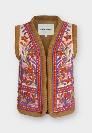 Femme portant une chemise blanche, un gilet brodé de fleurs, un jean bleu, des ballerines noires, tenant une pochette noire texturée, debout devant un fond blanc.