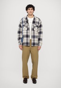 Plaid flannel shirt in white and dark purple, featuring two chest pockets. Paired with beige pants and dark loafers. Small blue bag slung across.