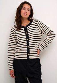 Cardigan cu dungi în nuanțe de crem și negru, având butoane aurii pe toată lungimea din față și o textură ribbată. Mâneci lungi și decolteu rotund.