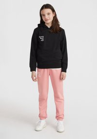 Zwarte hoodie met witte tekst op de voorkant, gecombineerd met lichtroze sweatpants, beide gemaakt van zachte stof; gedragen met witte sneakers.