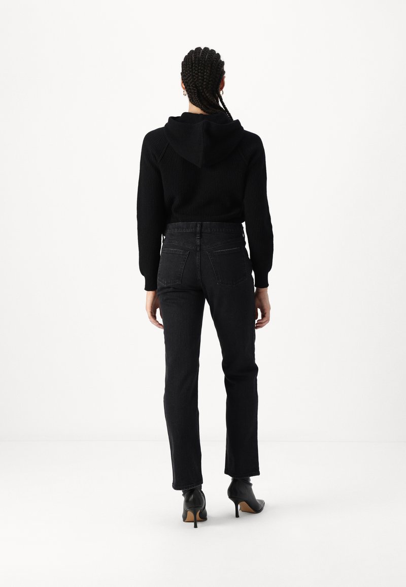 Schwarzer Rippstrickpullover mit Kapuze, kombiniert mit hochgeschnittenen schwarzen Jeans. Das Outfit hat gerade Beine und schwarze High Heels, von hinten betrachtet.