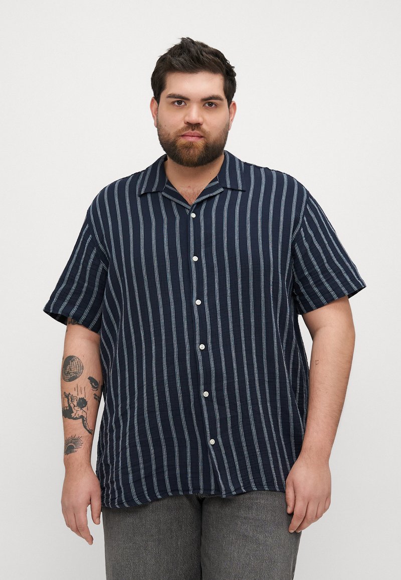 Camisa de botones a rayas marino con un corte relajado, mangas cortas y cuello tipo polo, que presenta rayas verticales en blanco y azul claro.