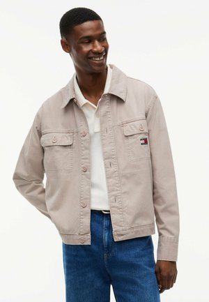 Lächelnder Mann mit einer beigefarbenen Jacke mit Knöpfen über einem weißen Poloshirt und blauen Jeans, steht mit einer Hand hinter dem Rücken.