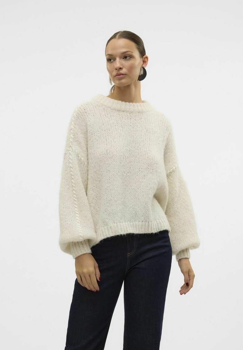 Pull en maille légère et texturée de couleur crème avec un large col ras du cou et des manches volumineuses. Associé à un denim foncé.