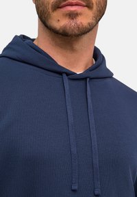 Uomo che indossa una felpa blu navy a coste con cappuccio e lacci, mostrando la parte inferiore del viso, il mento e il collo su sfondo bianco.