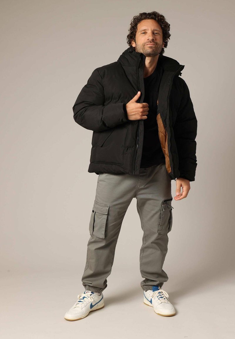 Veste rembourrée noire avec un col haut, dotée d'une doublure marron, associée à un pantalon cargo gris et des baskets blanches avec des accents bleus.