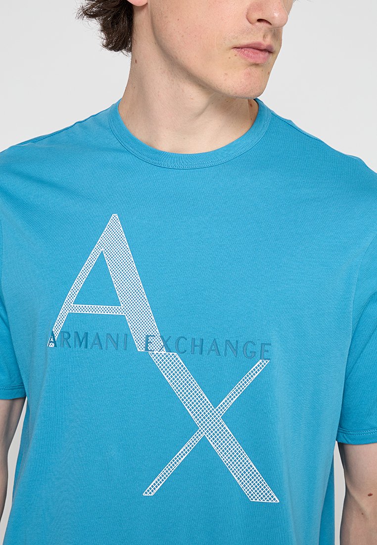 Giovane uomo che indossa una maglietta blu Armani Exchange con grandi lettere bianche "A X" e un testo della marca più piccolo sul petto.