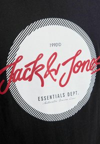 Zwarte T-shirt met een centraal witte cirkel met rode tekst "Jack & Jones" en gestreepte accenten. Bevat de tekst "1990©" en "ESSENTIALS DEPT."