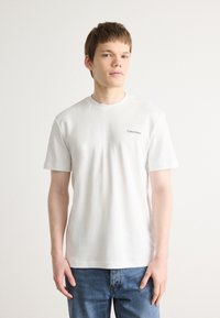 Fehér pamut póló rövid ujjal, kerek nyakkivágással és fekete "Calvin Klein" logóval a bal mellrészen. Kék farmernadrággal viselve.