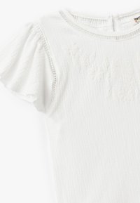 Top blanco de manga corta con un tejido texturizado y un diseño floral bordado. Presenta mangas con volantes y un escote redondo.