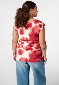 Rood en crèmekleurig gebloemd topje met korte kapmouwen en gelekte details in de taille, gedragen met lichtblauwe denim broek.