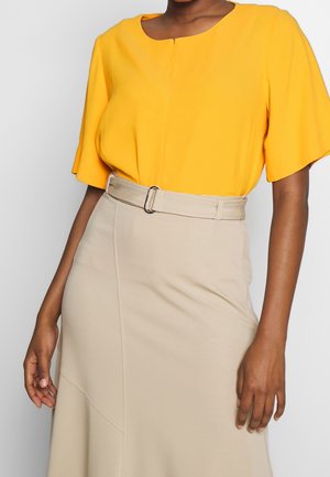 Frau trägt eine knallgelbe Kurzarmbluse, die in einen hoch taillierten beigen Rock mit passender Gürtelschnalle gesteckt ist.