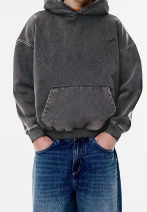 Person iført en falmet grå hættetrøje med forlommer og blå jeans, hænderne puttet i lommerne, stående mod en hvid baggrund.