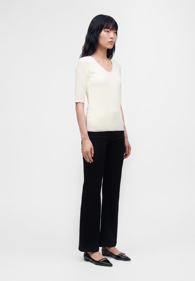 Femme aux longs cheveux noirs portant un haut blanc texturé à manches courtes, un pantalon droit noir et des ballerines pointues noires, debout de profil.