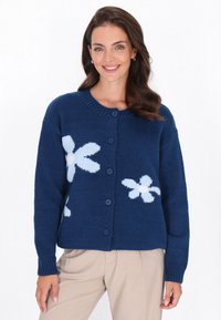 Cardigan lavorato a maglia blu navy con motivi floreali bianchi, chiusure con bottoni e polsini a coste. Il tessuto sembra morbido e texturizzato.