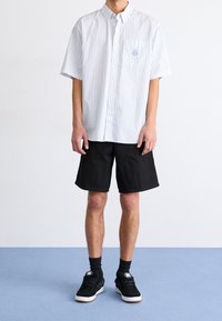 Lyhythihainen valkoinen raidallinen paita, jossa on rintatasku ja logo, yhdistettynä mustiin shortseihin ja mustiin tennareihin. Sileä kangas, rento istuvuus.