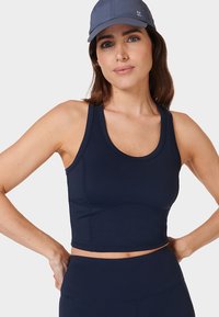 Vrouw in een marineblauwe sportcrop-top en legging met blauwe pet, staand tegen een effen lichte achtergrond, met handen op de heupen.