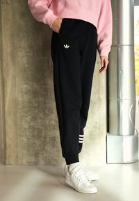 Osoba v černých teplácích Adidas se třemi bílými pruhy a logem, růžové mikině, bílých ponožkách a bílých teniskách, stojící uvnitř.