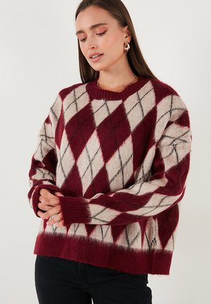 Vrouw in een bordeauxrood en crèmekleurig argyle patroontrui, met gouden oorbellen, staand tegen een effen lichte achtergrond met gevouwen handen.