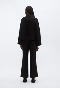 Kvinde med fletninger iført stor sort jakke, sorte bukser med vidde og sorte plateau sko, stående mod hvid baggrund.