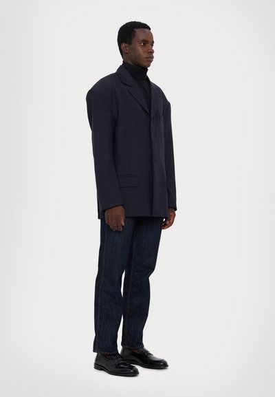 Übergroßer marineblauer Blazer mit strukturierten Schultern, einer einzelnen Brusttasche, kombiniert mit dunklen Jeans und schwarzen Lederschuhen.