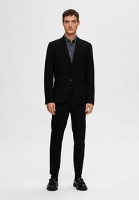 Črn smoking z oprijetim blazerjem, usklajenimi hlačami in strukturirano srajco z gumbki, ki vsebuje cvetlični vzorec; oblečen z črnimi čevlji.