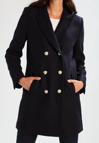 Marinblå dubbelknäppt kappa med guldknappar, långa ärmar och sidofickor; tillverkad i ett texturerat tyg.