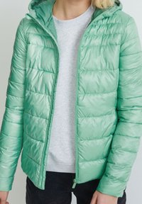 Chaqueta acolchada verde claro con capucha, que presenta una textura brillante, acolchado horizontal y cierre de cremallera. Lleva puesta una sudadera gris debajo.