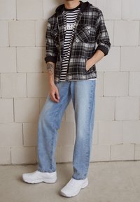 Giacca a camicia a quadri bianco e nero con cappuccio, T-shirt a righe bianco e nero sotto, jeans a gamba larga blu chiaro e sneaker bianche.