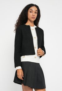Kvinde med langt krøllet hår iført en sort cropped jakke over en hvid sweater med gyldne hjerter og en mørk plisseret nederdel.
