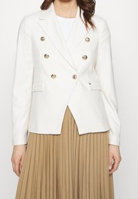 Donna che indossa un blazer doppiopetto bianco con bottoni dorati sopra una maglietta bianca e una gonna plissè beige, mostrata dalla nuca fino a metà coscia.